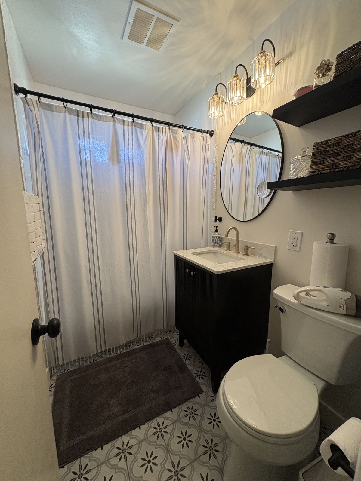 A finished bathroom with a dark vanity, a round mirror, wall shelving and patterned tile flooring.